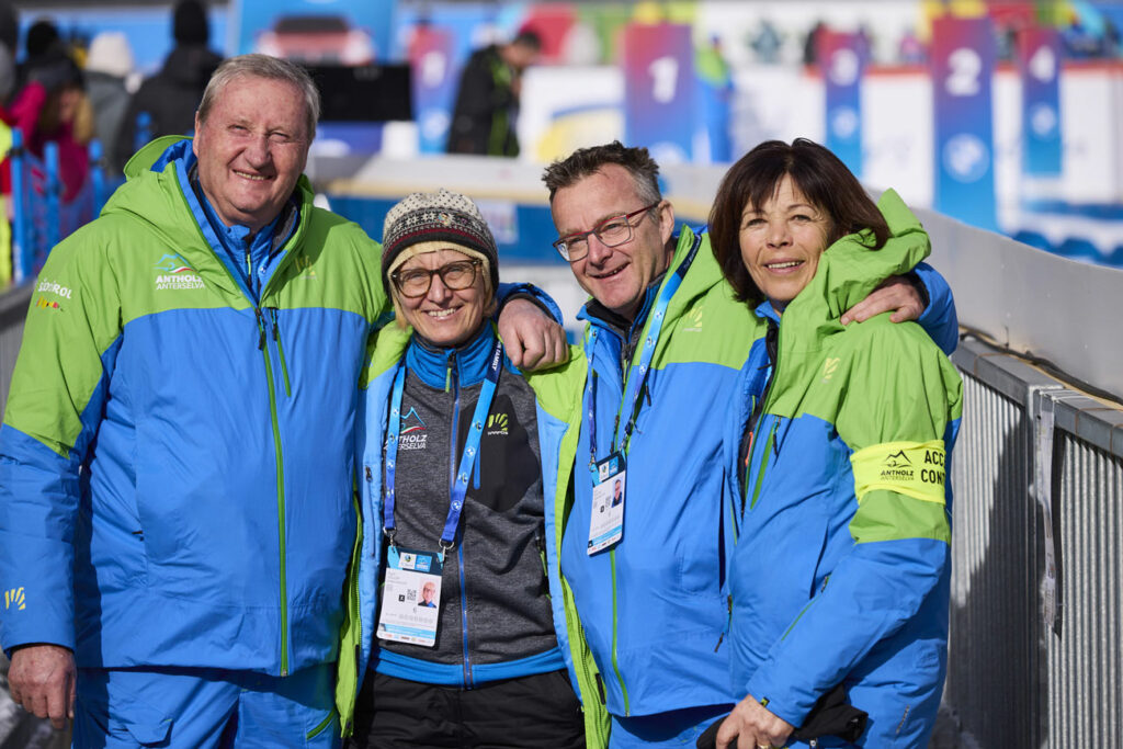 Vier freiwillige Helfer:innen des Biathlon-Wettbewerbs in Antholz stehen Arm in Arm und lächeln in die Kamera. Sie tragen blaue und grüne Teamjacken mit Logos von „Antholz Anterselva“ und Akkreditierungsausweise um den Hals. Im Hintergrund ist das Stadion mit farbigen Nummerntafeln zu erkennen.