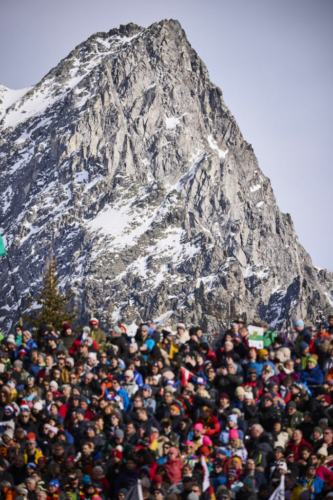 Im Vordergrund die voll besetzte Tribüne der Olympic Arena in Antholz, im Hintergrund die beeindruckende Bergkulisse