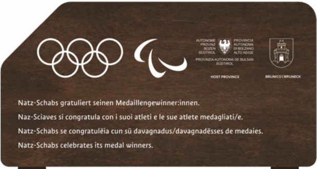 Ein braunes Schild mit den Symbolen der Olympischen und Paralympischen Spiele. Darunter stehen vier Gratulationssätze von Natz-Schabs an seine Medaillengewinner:innen in Deutsch, Italienisch, Ladinisch und Englisch. Oben rechts befinden sich die Logos der Autonomen Provinz Bozen und der Stadt Bruneck.